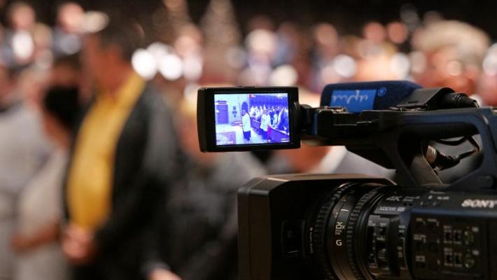 Eine professionelle Videokamera steht im Vordergrund und filmt eine Veranstaltung. Auf dem aufgeklappten Kameradisplay ist eine Gruppe von Menschen in einem Innenraum zu sehen. Im unscharfen Hintergrund befinden sich weitere Personen, die offenbar an der Veranstaltung teilnehmen.