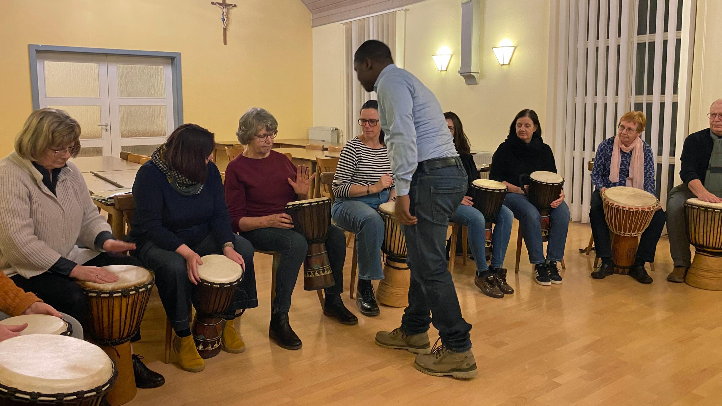 Eine Gruppe von Menschen sitzt in einem hell erleuchteten Raum in einem Halbkreis, jeweils mit einer Djembe‑Trommel vor sich. Ein stehender Anleiter in der Mitte gibt Hinweise oder führt etwas vor. Der Raum hat helle Holzböden, hohe Fenster mit Lamellen und warmgelbe Wände mit Wandlampen. Alle wirken konzentriert und in eine gemeinschaftliche Trommelübung vertieft.
