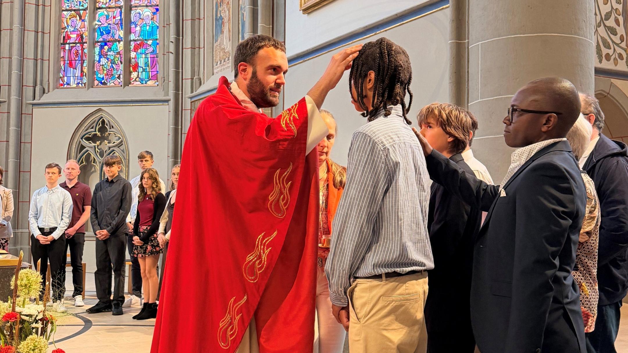 Ein Priester in einem roten Gewand mal mit seinem Finger ein Kreuzzeichen aus Öl auf die Stirn eines Jugendlichen. Hinter ihm steht sein Pate und legt seine Hand auf die Schulter des Jungen.