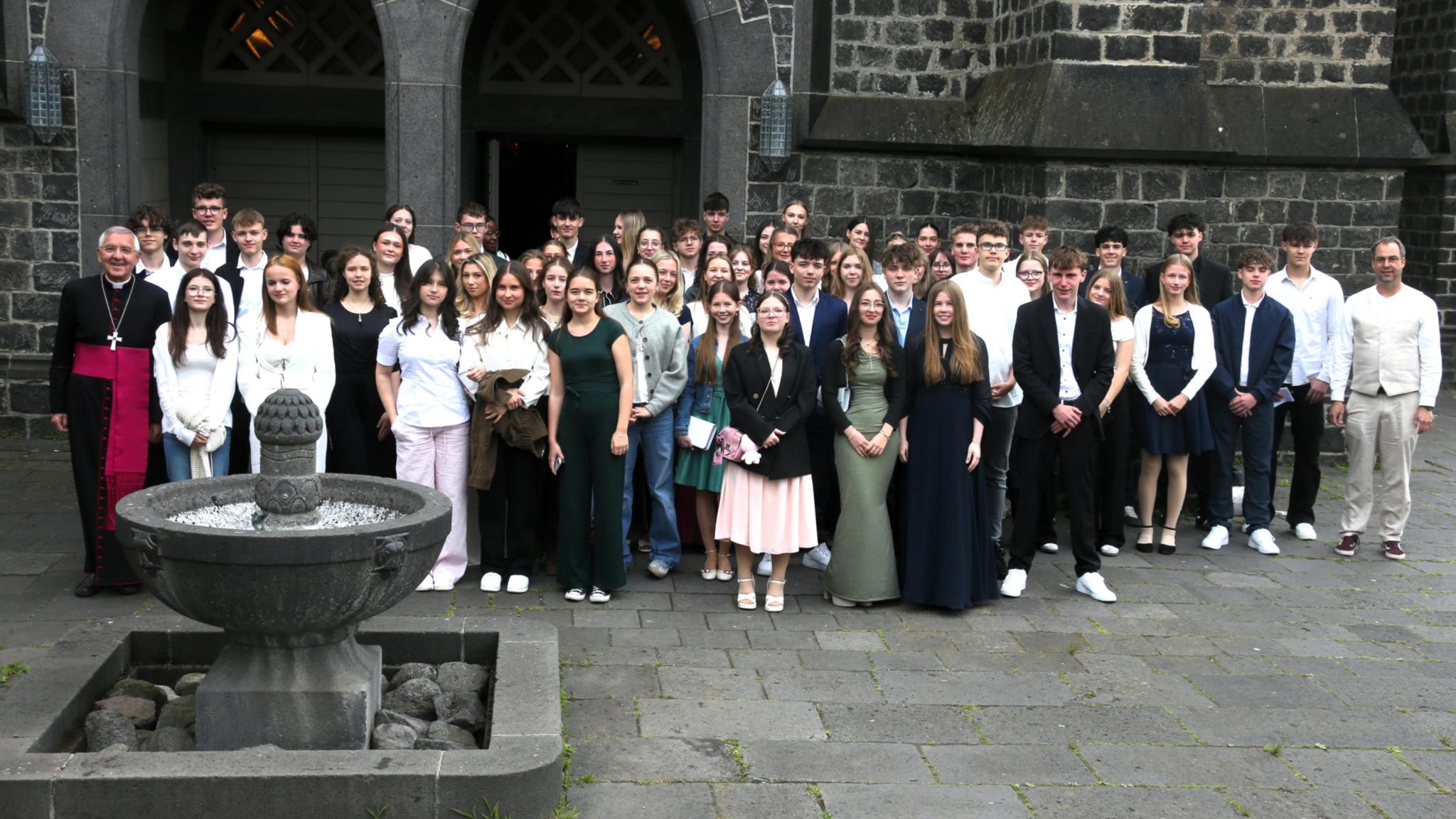 Gruppenfoto mit vielen jungen Menschen vor einer Kirche mit dunklen Steinen.