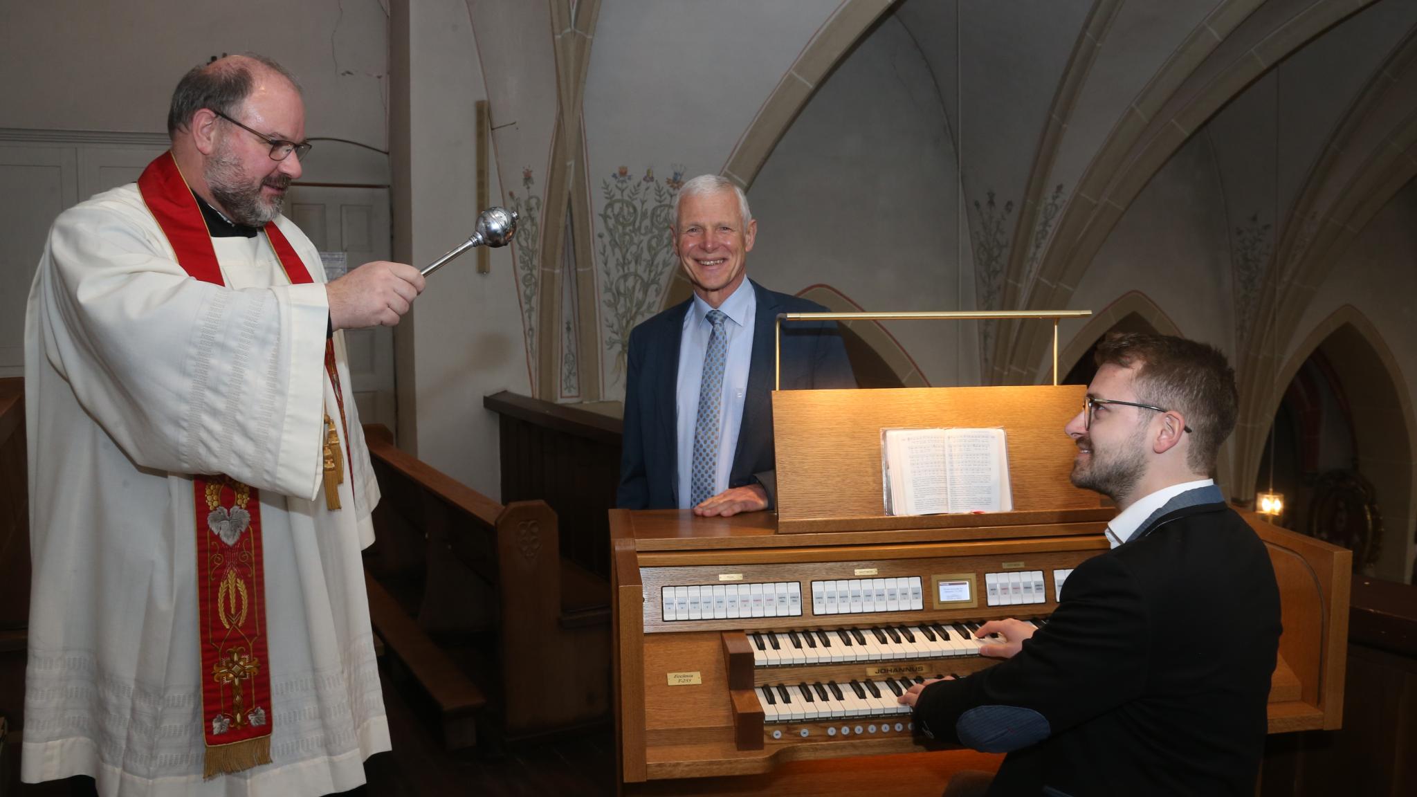 Ein Pfarrer besprengt eine Orgel mit Weihwasser, ein Mann sitzt an der Orgel, ein weiterer Mann steht hinter der Orgel und lächelt.