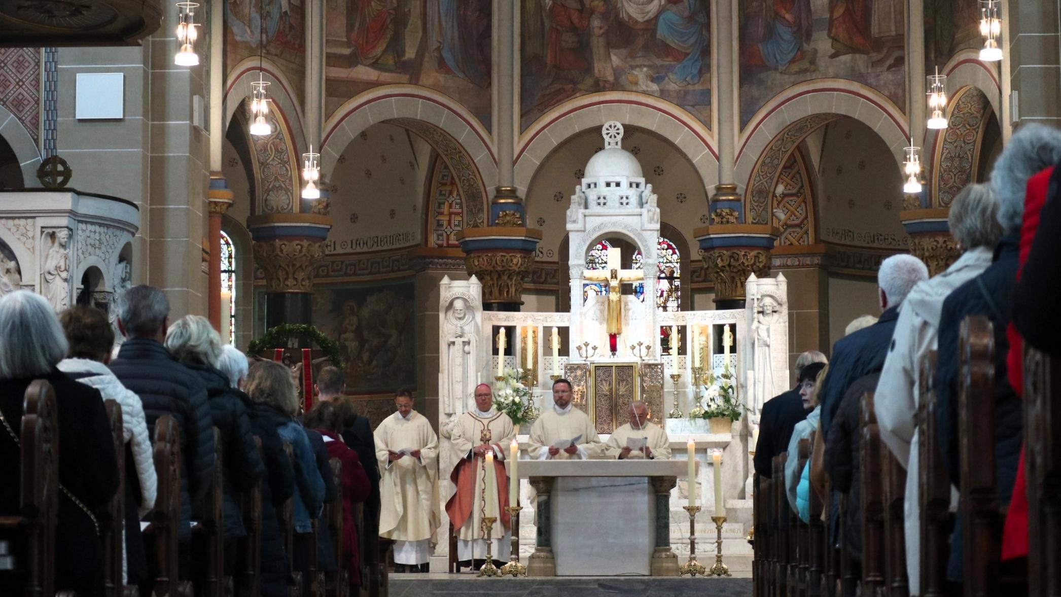 Priester stehen an einem weißen Altar, zu sehen ist auch ein langer Mittelgang mit vielen Personen, die in Kirchenbänken sitzen.
